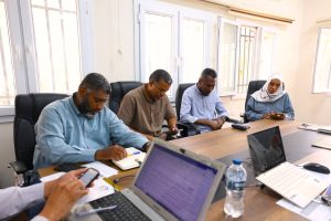 Five professionals seated around a conference table, taking notes and using laptops/tablets in a bright meeting room.