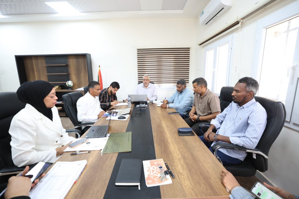 Eight professionals seated around a long conference table in a bright office, with laptops, papers, and pens as they discuss matters from documents and notebooks this meeting room setup shows teamwork and collaboration.