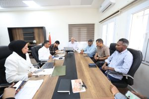 Eight professionals seated around a long conference table in a bright office, with laptops, papers, and pens as they discuss matters from documents and notebooks this meeting room setup shows teamwork and collaboration.