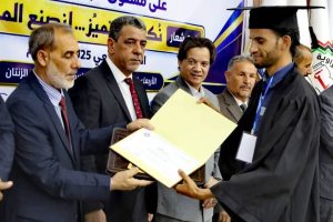 Graduate in cap and gown receives a certificate from a man in a suit at a formal graduation ceremony, with onlookers and a banner in Arabic behind them.