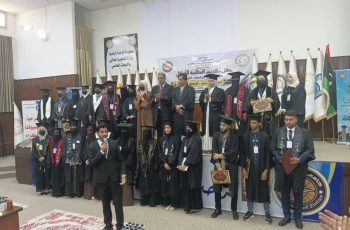 Group of graduates in caps and gowns standing on a stage during a university graduation ceremony, banners and flags in the background.