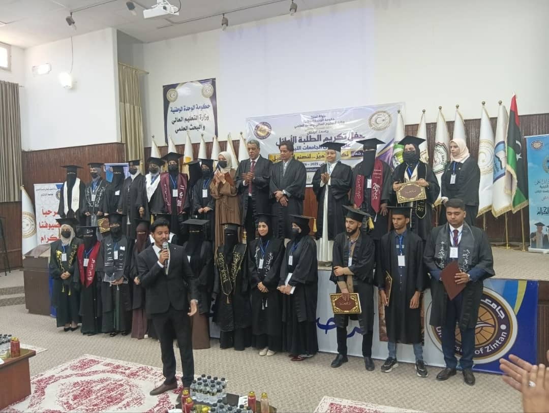 Group of graduates in caps and gowns standing on a stage during a university graduation ceremony, banners and flags in the background.