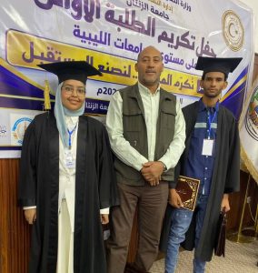 Three people pose at a graduation ceremony: two graduates in caps and gowns (woman left in hijab and glasses, man right holding a diploma) with a man in a vest in the middle; large Arabic banner in the background.
