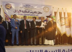 Group of men on a stage during an Arabic ceremony; a graduate in a robe and mortarboard hands a certificate to another man, with flags and a banner in the background.