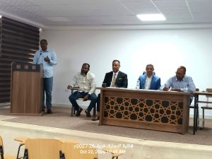A man stands at a wooden podium with a microphone, speaking to a panel of four men seated at a long desk in a conference room.