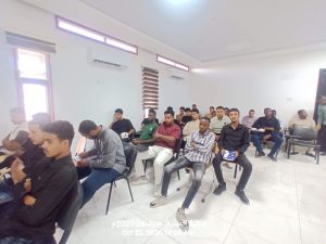 IMG-20261024-WA0007 - جامعة سبها | Sebha University A classroom filled with students seated in rows, listening to a lecture in a bright white room with large windows on the left.