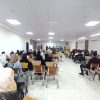 Crowded conference room with a panel of speakers at the front and attendees seated in rows.