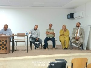 IMG-20261024-WA0010 - جامعة سبها | Sebha University Five professionals seated in a panel discussion; leftmost person writes at a desk while others listen in a bright room.