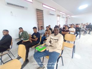 IMG-20261024-WA0011 - جامعة سبها | Sebha University A classroom full of students seated in rows, looking toward the front, with notebooks and pens on their desks and a bright, airy room behind them.