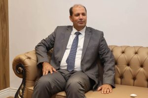 IMG-20261025-WA0022 - جامعة سبها | Sebha University Middle-aged man in a gray suit and polka-dot tie sits on a tan tufted sofa in a neutral room.