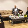 Three men in suits seated on beige tufted sofas around a coffee table in a formal business meeting room.