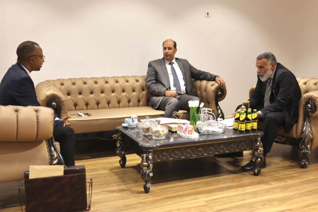 IMG-20261025-WA0023 - جامعة سبها | Sebha University Three men in suits seated on beige tufted sofas around a coffee table in a formal business meeting room.