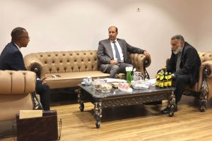 IMG-20261025-WA0023 - جامعة سبها | Sebha University Three men in suits seated on beige tufted sofas around a coffee table in a formal business meeting room.
