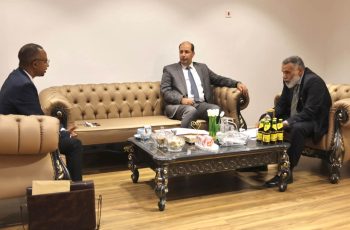 Three men in suits seated on beige tufted sofas around a coffee table in a formal business meeting room.