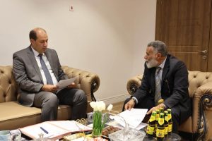 IMG-20261025-WA0024 - جامعة سبها | Sebha University Two men in suits sit on a beige leather sofa in a meeting, reviewing documents on a coffee-table full of papers and drinks.