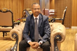 IMG-20261025-WA0025 - جامعة سبها | Sebha University Man in a navy suit and striped tie sits on a beige tufted sofa in a wood-paneled, formal office.