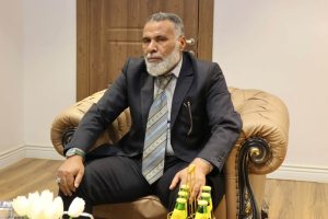 IMG-20261025-WA0026 - جامعة سبها | Sebha University Older man with a gray beard in a dark pinstripe suit sits on a tan leather armchair in an office, hands resting on his knees.