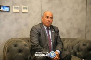 Man in a dark suit and red tie sits on a tufted gray sofa in a neutral office space, with the University of Benghazi logo visible at the bottom.