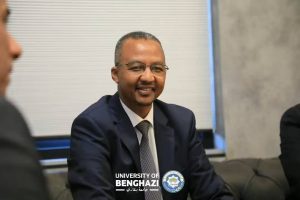 Portrait of a smiling man in a dark suit and tie at a meeting, with the University of Benghazi logo visible on his lapel/foreground.