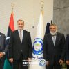 Four men in suits pose for a formal photo in front of Libyan flags and a university banner, at University of Benghazi events.