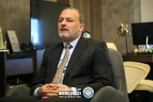 Man in a dark pinstriped suit and patterned tie sits in a modern office, hands clasped, with the University of Benghazi logo visible at the bottom.