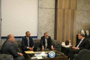 Four men in suits sit on a tufted gray sofa in a modern office lounge, discussing; Benghazi emblem on the table.