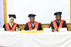 Three graduates in black gowns and mortarboard caps sit at a panel table during a ceremony, with name placards and drinks in front of them