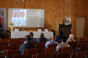 Speaker at podium in a wood-paneled room presenting a slide showing kidneys and Arabic text to an audience of seated attendees.