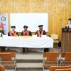 Panel of three graduates in red gowns and mortarboards seated at a table on a stage, with a speaker at a podium to the right in a wood-paneled room.