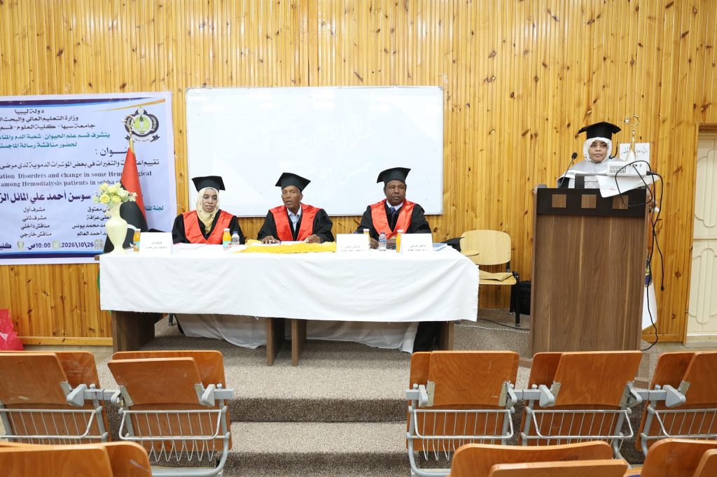 IMG-20261026-WA0016 - جامعة سبها | Sebha University Panel of three graduates in red gowns and mortarboards seated at a table on a stage, with a speaker at a podium to the right in a wood-paneled room.