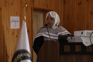 Woman in a hijab speaks at a podium with a microphone in front of wood-paneled walls.