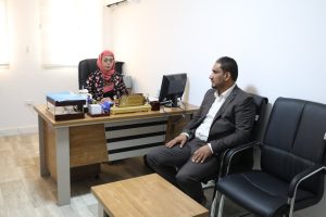 IMG-20261026-WA0025 - جامعة سبها | Sebha University Woman in a pink hijab sits behind a wooden desk with a computer and office supplies, facing a man in a suit seated in a black chair across from her in an office.