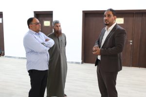 IMG-20261026-WA0036 - جامعة سبها | Sebha University Three men standing in a bright hallway, one in a suit speaking while the other two listen attentively.