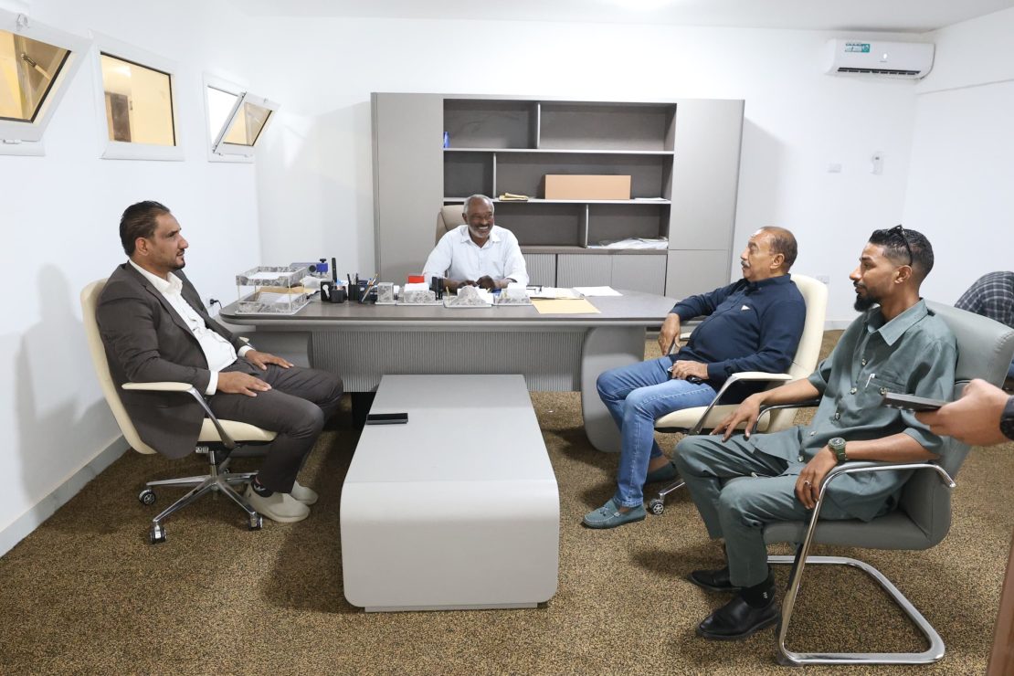 Five men sit around a grey conference desk in a bright office, chatting during a meeting.