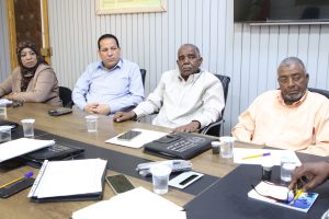 Four people sit at a conference table with papers, notebooks, and cups in a meeting room.