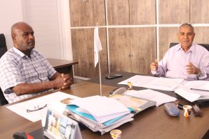 Two men in a meeting room sit across a cluttered conference table with papers and cups nearby.