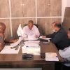 Four men sit around a conference table reviewing documents in a tidy office; a small flag stands near the center.