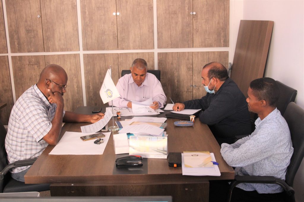 IMG-20261027-WA0018 - جامعة سبها | Sebha University Four men sit around a conference table reviewing documents in a tidy office; a small flag stands near the center.