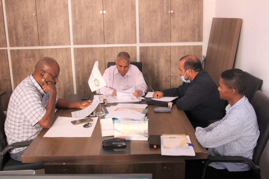 Four men sit around a conference table reviewing documents in a tidy office; a small flag stands near the center.