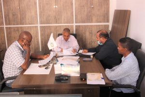 Four men sit around a conference table reviewing documents in a tidy office; a small flag stands near the center.