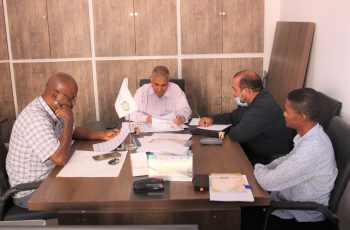 Four men sit around a conference table reviewing documents in a tidy office; a small flag stands near the center.