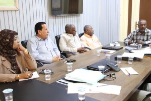 Six adults seated around a conference table with papers, cups, and phones, listening in a meeting.