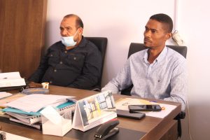 Two men sit at a cluttered office desk; one wears a dark shirt and a surgical mask, the other wears a light patterned shirt as they talk. Paperwork, notebooks, and a calendar are spread across the desk.