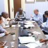 Six men seated around a conference table discussing documents and taking notes, with cups and phones on the table.