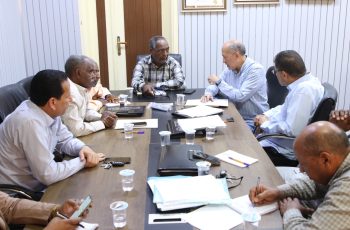 Six men seated around a conference table discussing documents and taking notes, with cups and phones on the table.