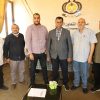 Six men stand together in an office, posing in front of a wall with a crest and Arabic text; a glass coffee table with papers and boxes sits nearby.