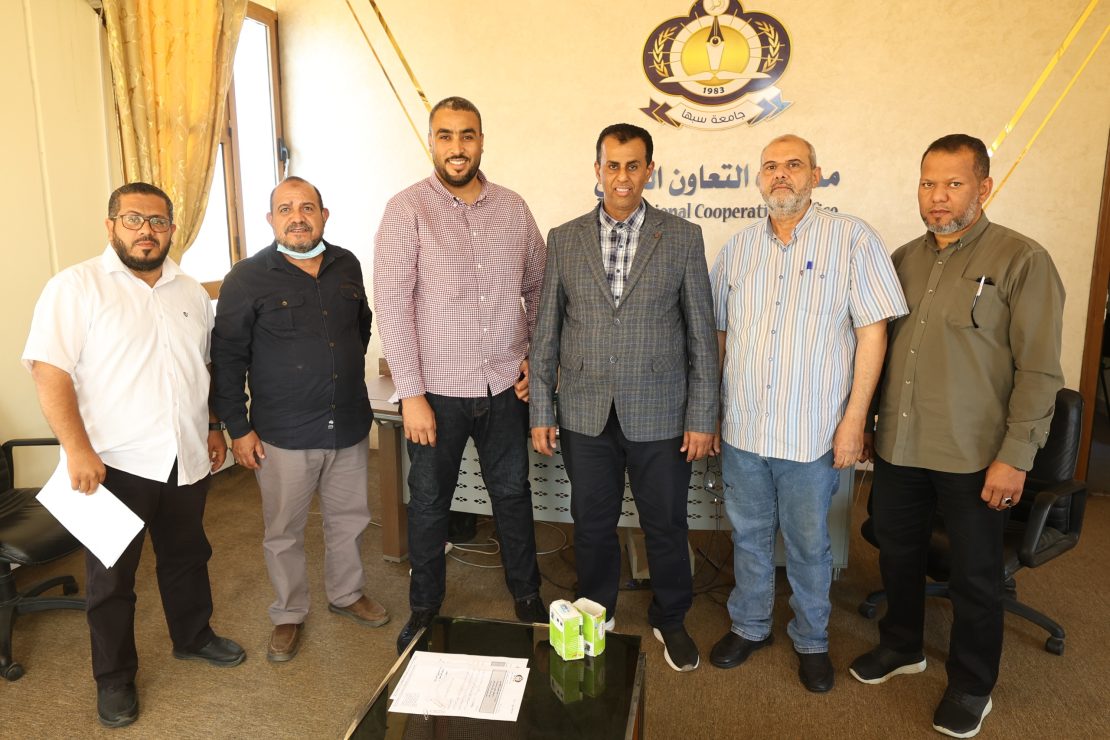 Six men stand together in an office, posing in front of a wall with a crest and Arabic text; a glass coffee table with papers and boxes sits nearby.