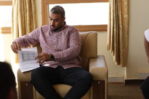 IMG-20261028-WA0016 - جامعة سبها | Sebha University Man in a red checkered shirt sits in a beige armchair, reading and marking papers with a pencil in hand.