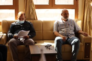 IMG-20261028-WA0018 - جامعة سبها | Sebha University Two men sit on a beige sofa by sunny windows; one wearing a mask and reading papers, the other gazing to the side in a striped shirt.