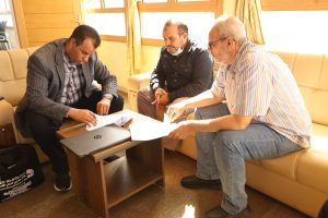 IMG-20261028-WA0019 - جامعة سبها | Sebha University Three men sit on a beige sofa around a wooden table, reviewing documents together in a bright room with a laptop nearby.